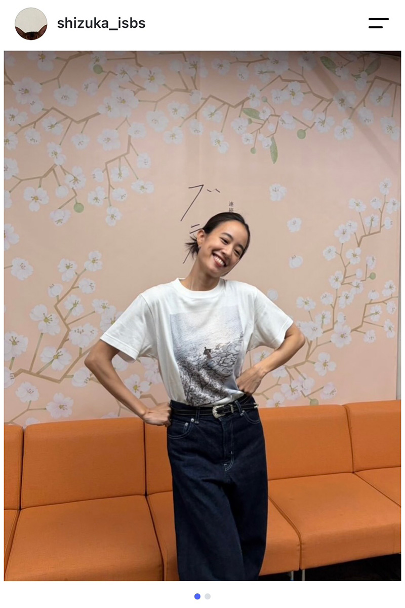 ブラッサムの白い番組Tシャツと濃紺デニムを着て、桜の絵の前で笑う石橋静河さん