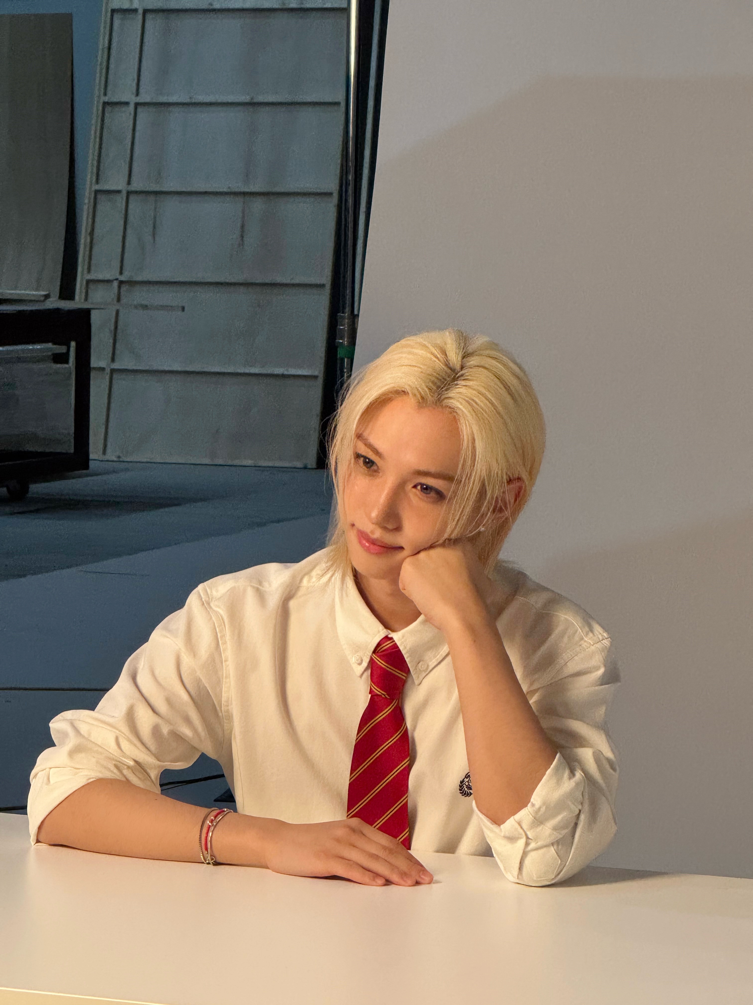 白シャツと赤いネクタイでテーブルに座り、頬に手を添えるStray Kids・FELIXさん