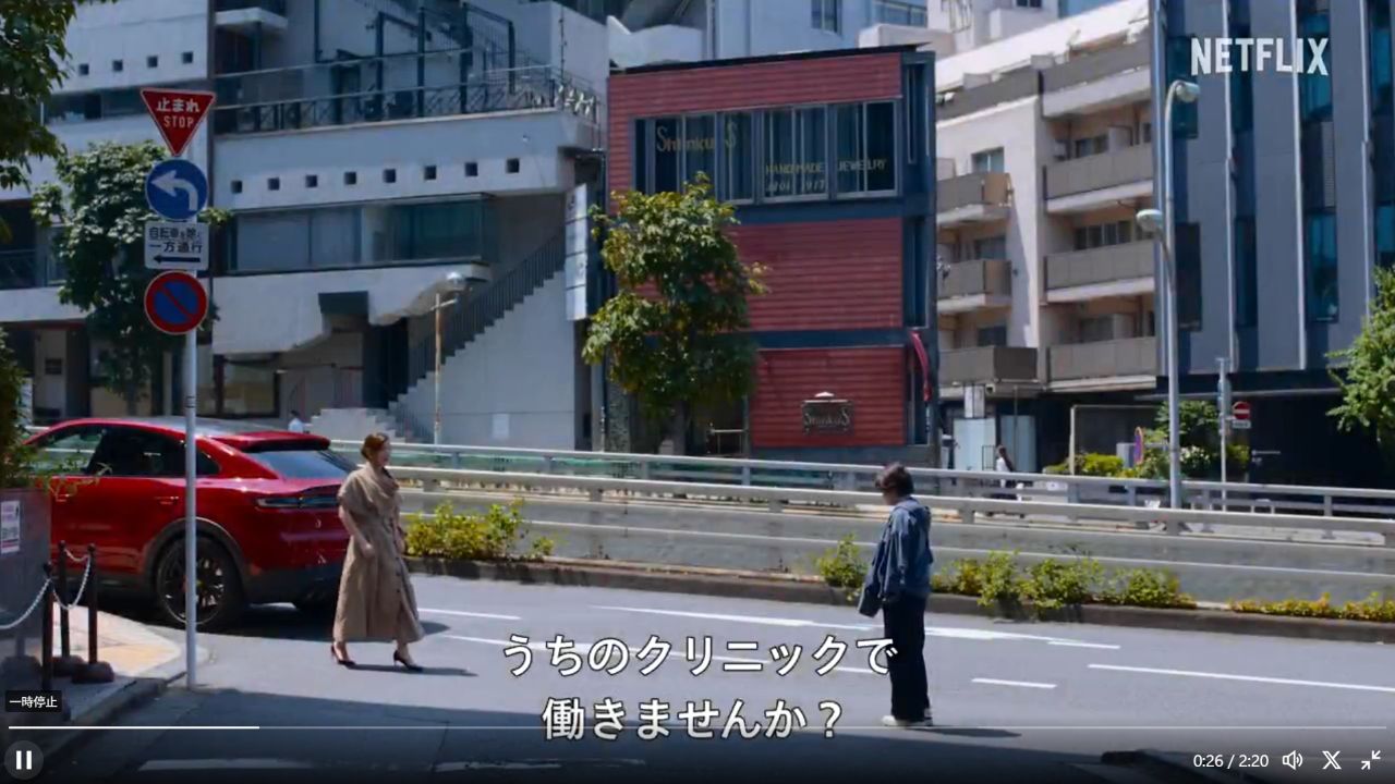 公式予告のスカウト場面で、赤い車のそばに立つ仲里依紗さん。ベージュのコートの襟、短い袖、ベルトと長い丈が見える