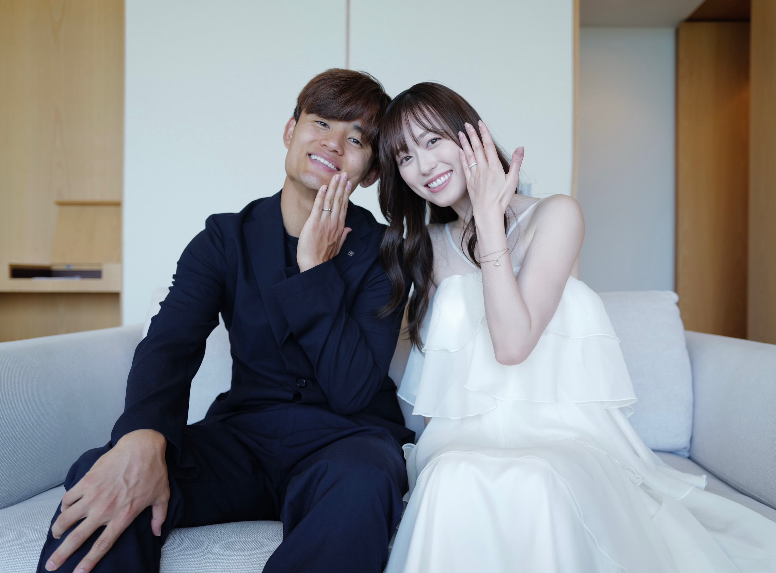 濃紺のスーツに花の襟ピンを付けた久保建英さんと、隣で白いドレスを着る福原遥さん