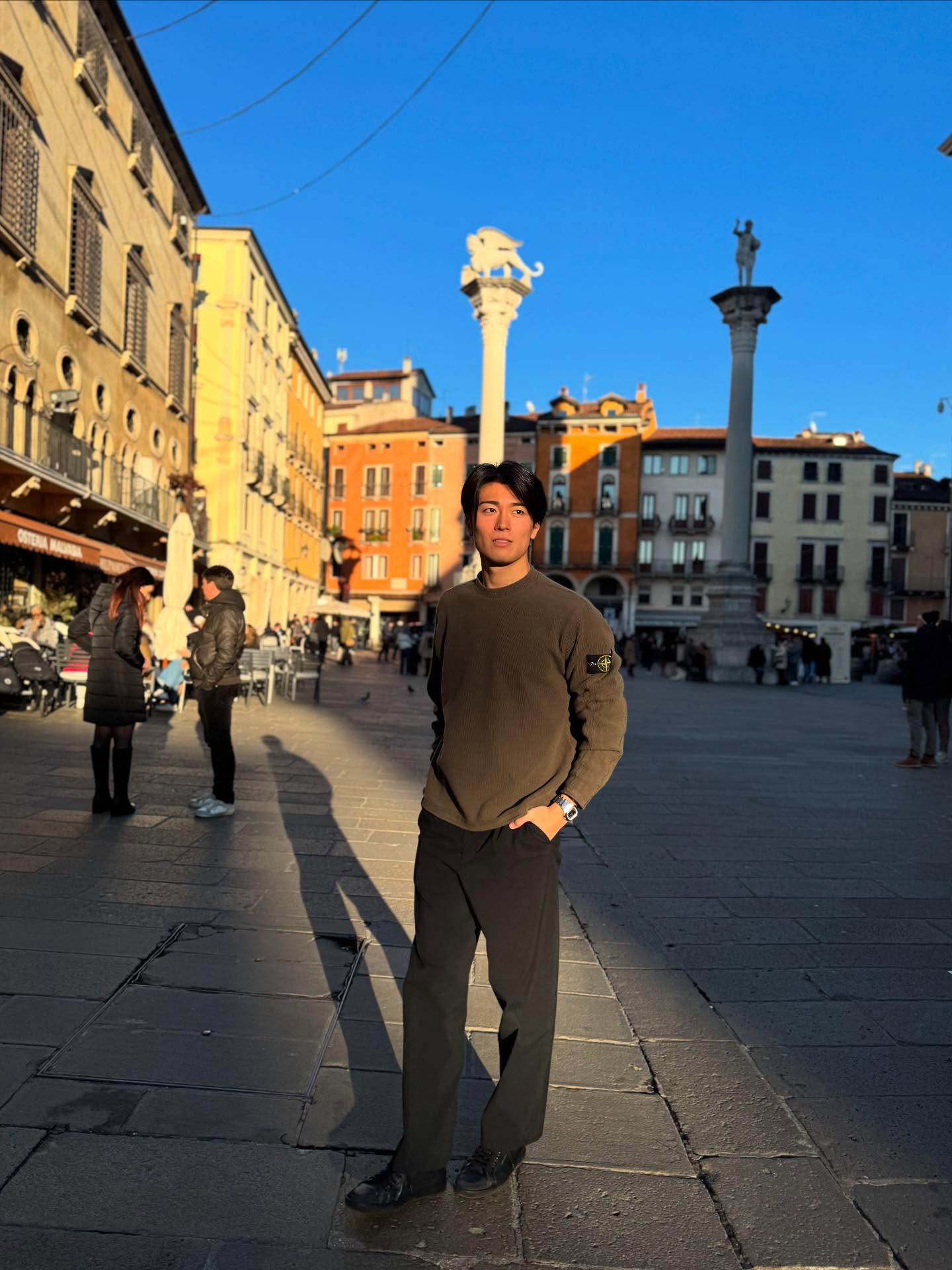 イタリアの広場で、オリーブ色のリブニットと黒いパンツを着た中村敬斗さん