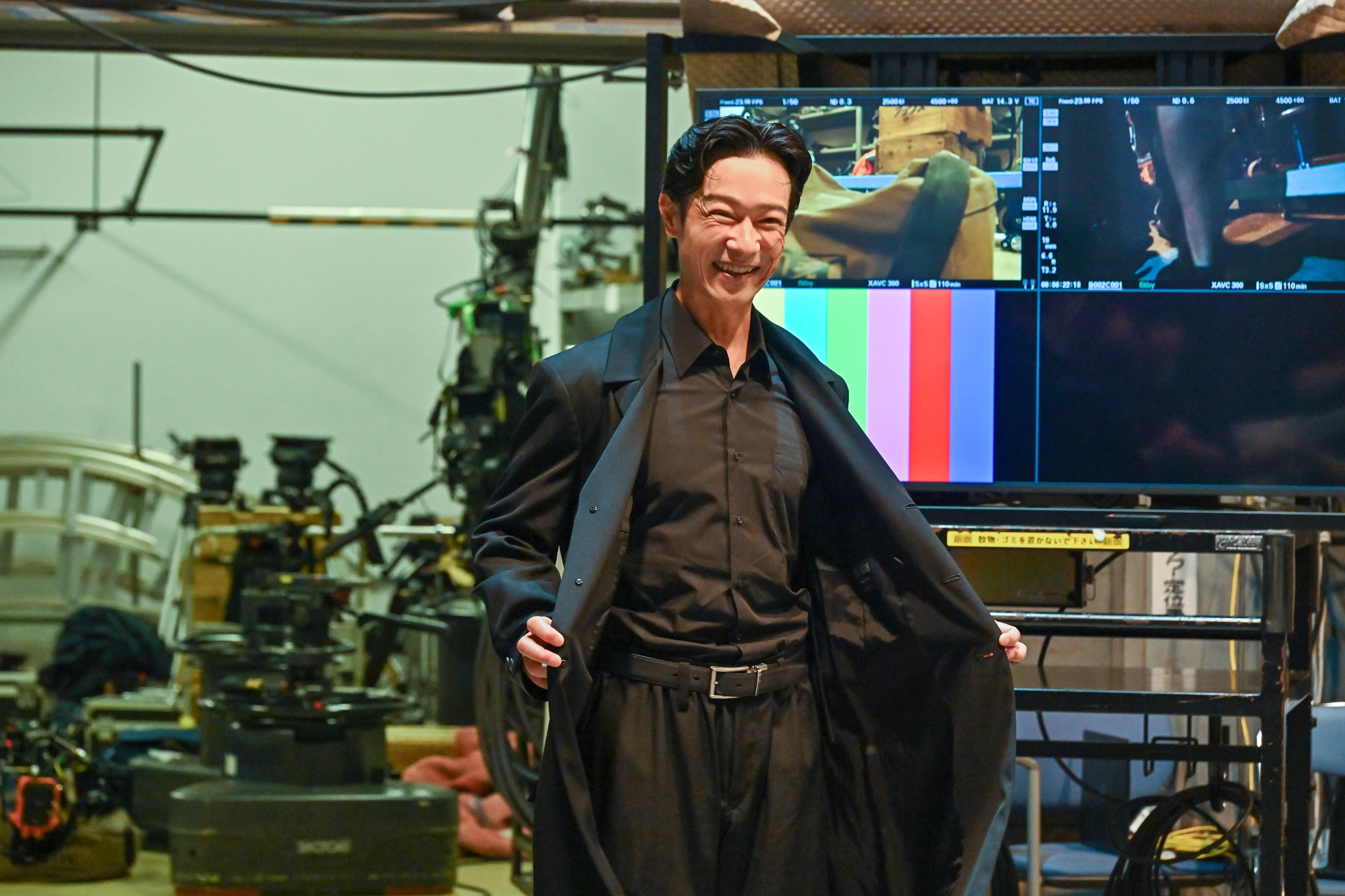 VIVANTの撮影オフショットで、コートを広げて黒いシャツを見せながら笑う堺雅人さん