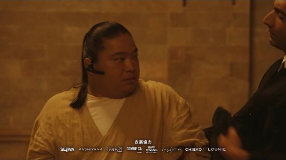 『VIVANT』第20話、1分22秒のドラム。左寄りの富栄ドラムさんは、前を重ねた襟のベージュの上衣と白いインナーを着ている